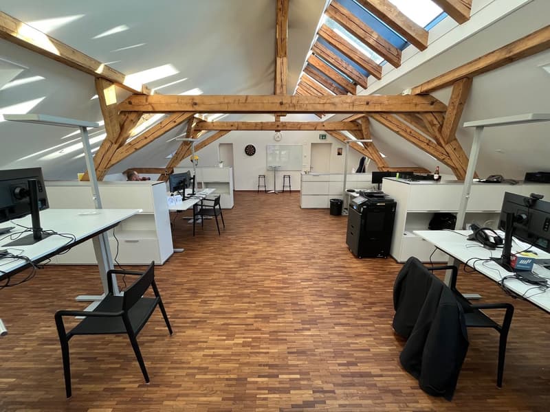 Open office space with wooden beams, wooden floors, desks, chairs, monitors, whiteboard, clock, trash bin, printer, storage cabinets, and a clock on the wall.