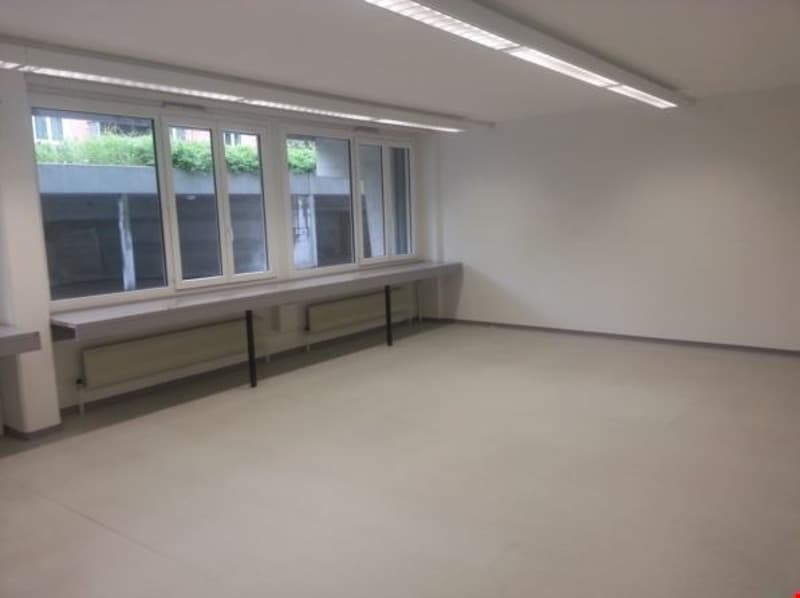 Empty room with white walls, large windows, gray floor, and a long bench with radiators