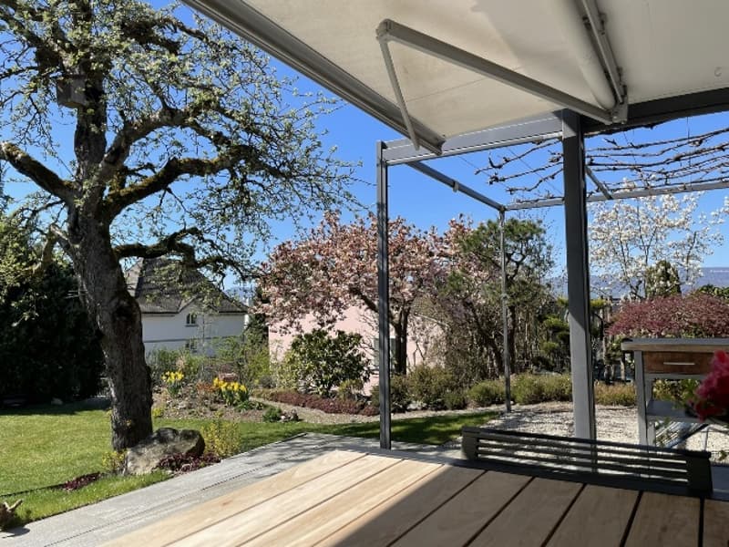 Wooden deck, retractable awning, garden with trees and flowers, house in the background