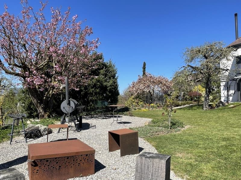 Outdoor seating area with tables and chairs, gravel pathway, trees, and a house in the background