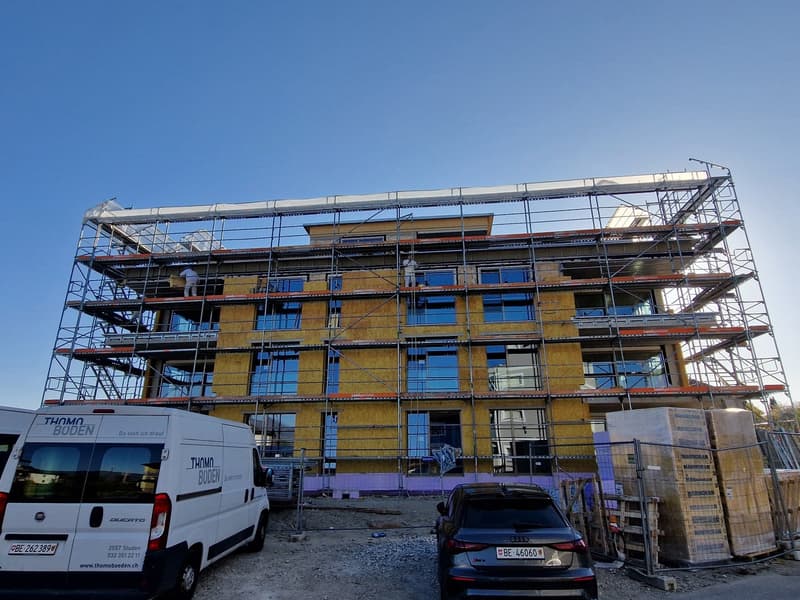 A multi-story building under construction, with scaffolding surrounding the exterior. There are construction vehicles, including a van and an Audi car, parked in front of the building. The building appears to be made of yellow brick or concrete material.