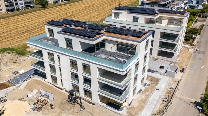 Multi-story apartment building with white exterior, large windows, balconies, and solar panels on the roof. There is a construction site in the foreground with equipment and materials.