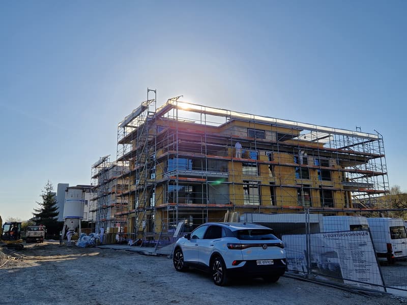 A multi-story building under construction, surrounded by scaffolding and construction materials. A white Volkswagen SUV is parked in front of the building.
