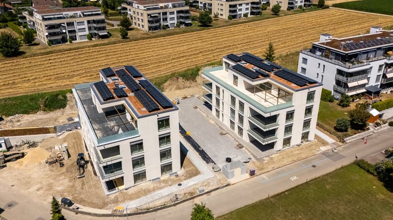 Multi-story apartment buildings with solar panels on the roofs, surrounded by a grassy field. There appears to be a parking area or driveway in front of the buildings.