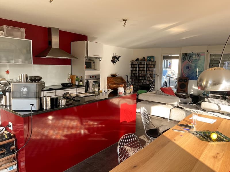 Modern kitchen with red walls, stainless steel appliances, modern kitchen island, black countertop, dining table, white and black chairs, living room area with grey sofa and coffee table