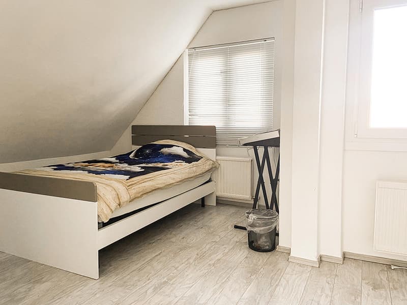 Single bed, white bed frame, white walls, wooden floor, window with blinds, radiator, trash can