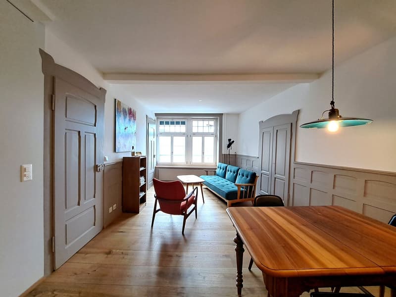 Empty room with wooden floor, white walls, wooden table, red chair, blue couch, wooden cabinet, window, lamp