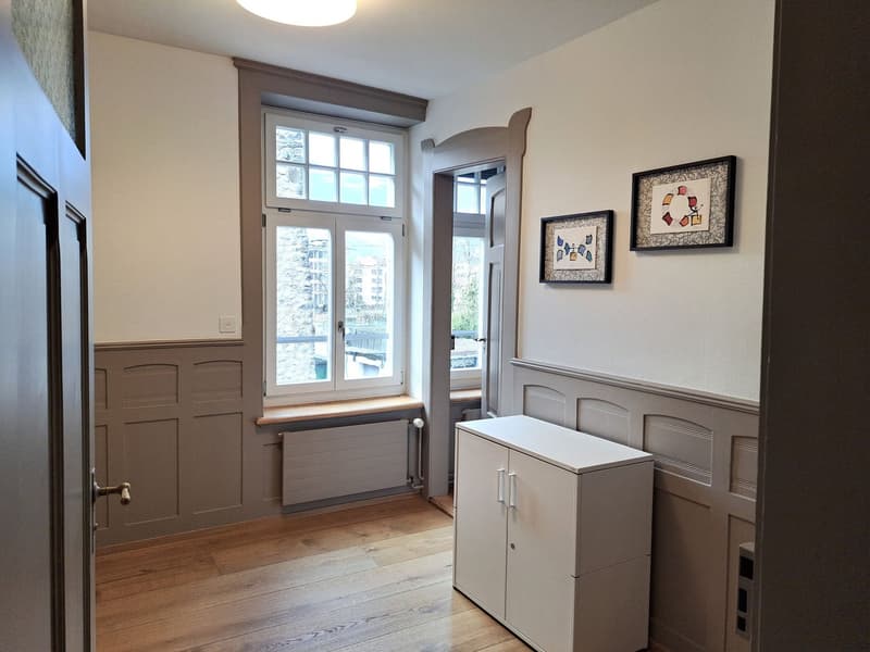 A hallway with wooden floor, white walls, a white cabinet, a window with a bench, and two framed pictures on the wall.