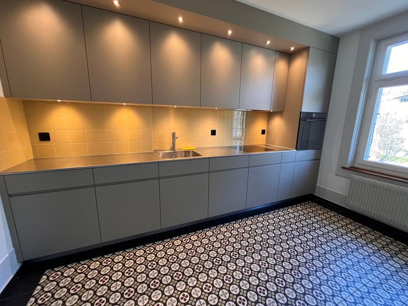 modern kitchen with gray cabinets, stainless steel countertops, tiled backsplash, under-cabinet lighting, double sink, faucet, oven, window with view