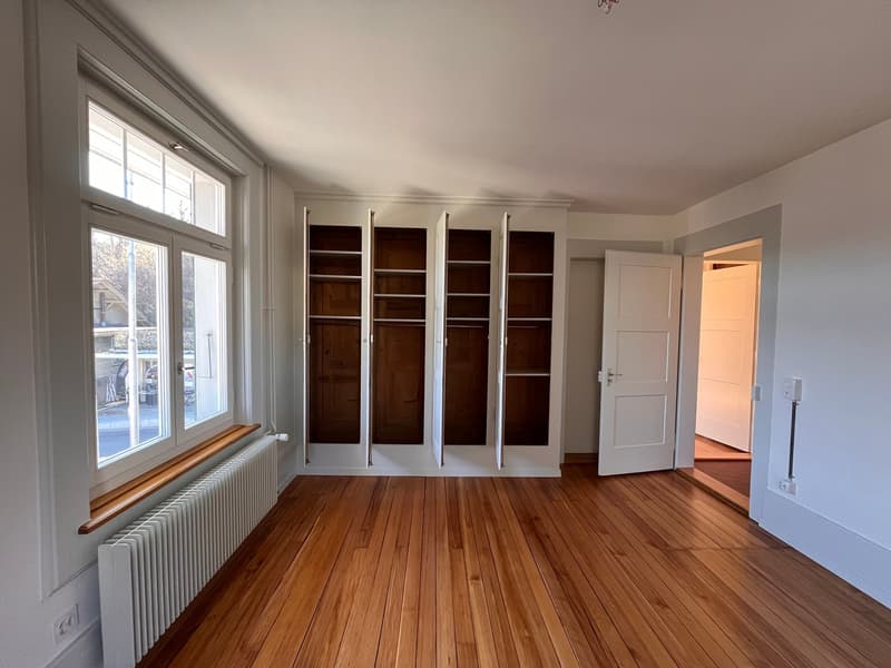 Empty room with wooden floor, white walls, large window, built-in wardrobes, radiator