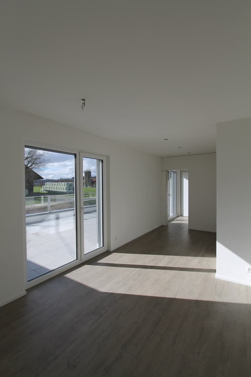 Spacious living room with large windows, hardwood flooring, and access to a balcony or terrace overlooking the outdoor area.
