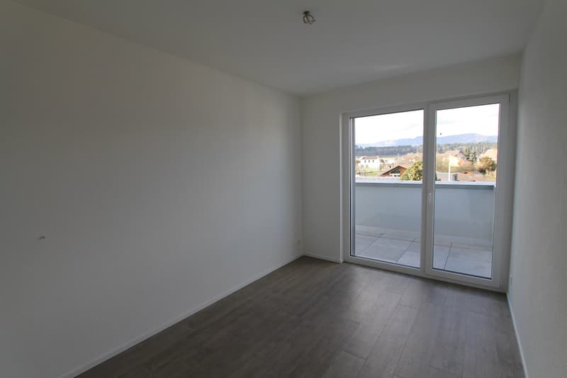 Spacious bedroom with large windows providing a view of the surrounding area, hardwood flooring, and a balcony or terrace accessible through the sliding glass doors.