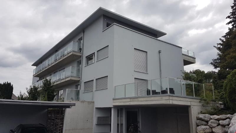3-story modern apartment building with gray exterior, balconies, and a parking area on the ground level