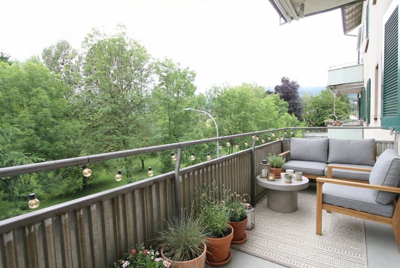 Outdoor area with a seating arrangement featuring a small table, two grey couches, string lights, a decorative lantern, and potted plants.