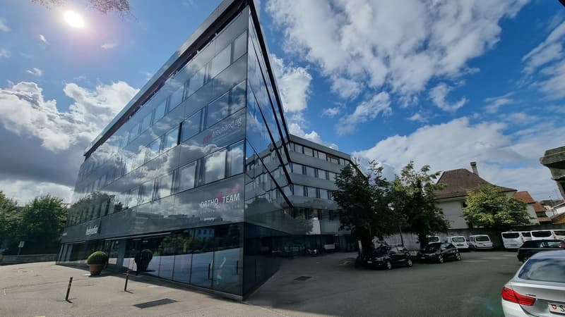 Modern office building with glass facade, multiple floors, parking lot in front, trees and plants around