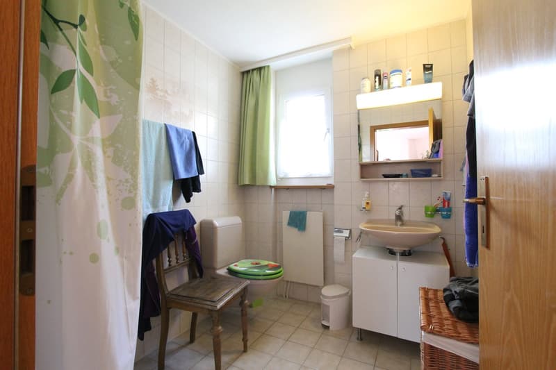 Bathroom with toilet, sink, mirror, and storage cabinet. A wooden chair and a basket are also present.