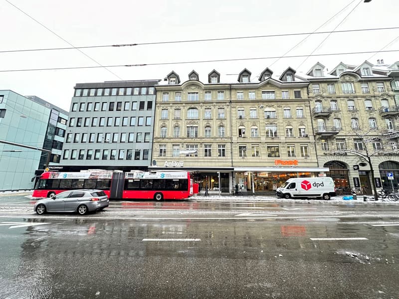 Multiple buildings in city. Large building in front with sign 'Fust' and smaller building behind it with sign 'Artiker'. Cars driving by on a road. Bicycles parked in front of building.