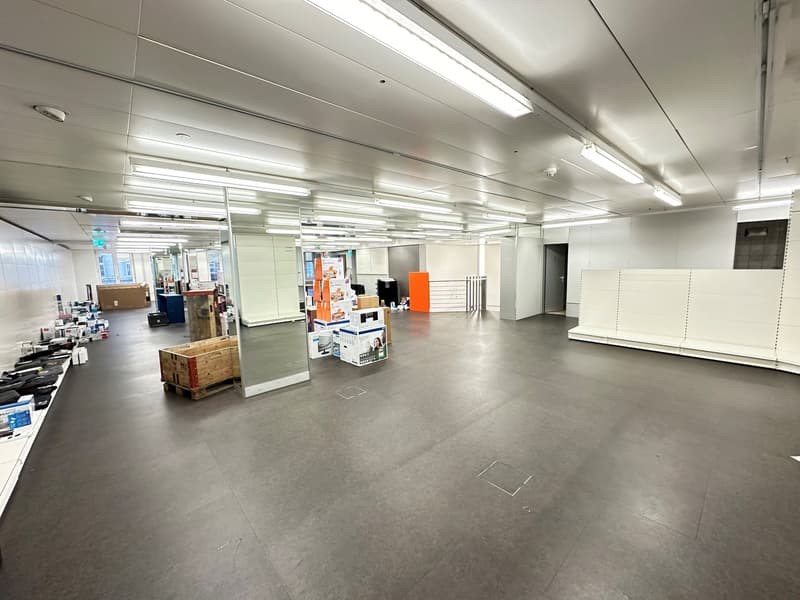 A wide open space with multiple fluorescent lights installed on the ceiling. There are shelves with some storage boxes on the left side of the room. Some storage boxes are also placed on the floor.