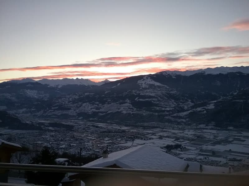 Overlook of a village from a balcony, snowy mountains and sunset in the background.