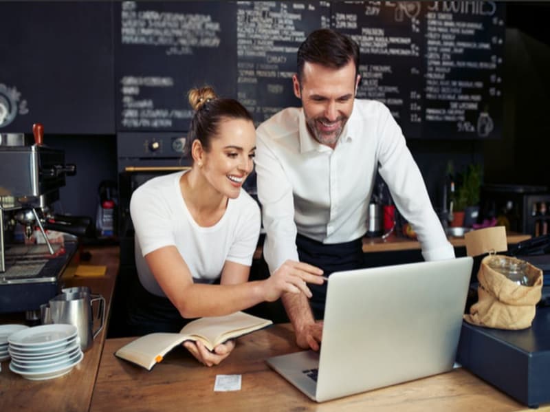 The image shows a couple working together at a cafe or restaurant. They are standing at a counter, with a laptop computer and a notebook in front of them. The background features a chalkboard menu and other cafe decor. The couple appear to be engaged in a