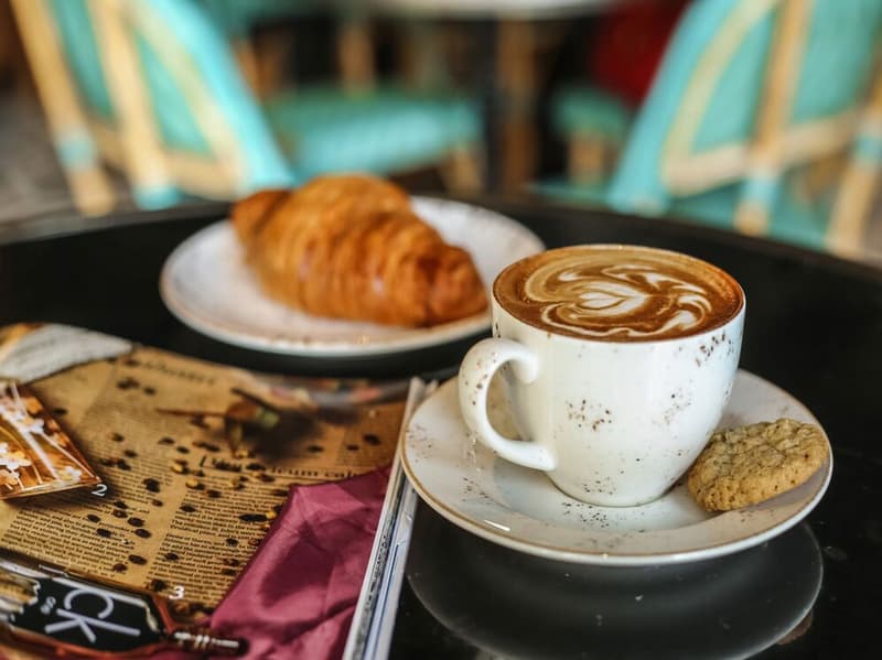 A cup of coffee with a latte art design, served with a croissant on a plate. The setting includes a newspaper and other items on the table, suggesting a cozy cafe environment.