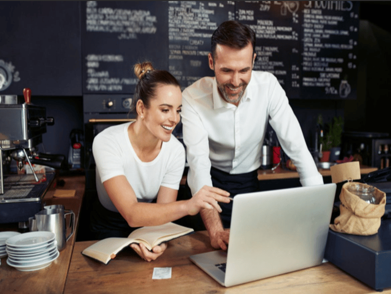 The image shows a couple working together at a cafe or restaurant. They are sitting at a counter, with a laptop and a book open in front of them. The background includes a chalkboard menu and other cafe equipment, suggesting this is a commercial food serv