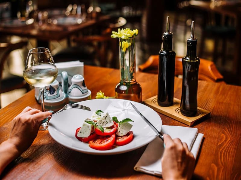 A person setting a place at a wooden table with a white plate of tomatoes and mozzarella, a wine glass, and two oil bottles on a wooden stand.