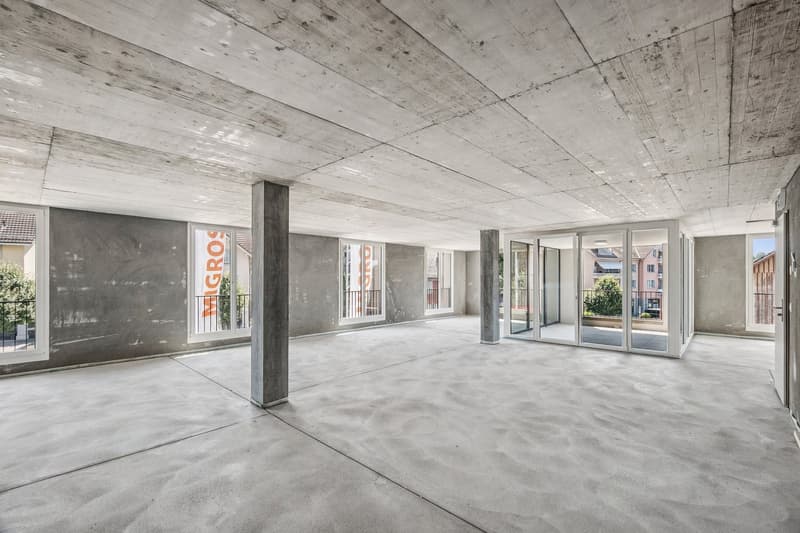 Large open-plan space with concrete ceiling and floor, glass sliding doors leading to a balcony or terrace, and exposed concrete columns.