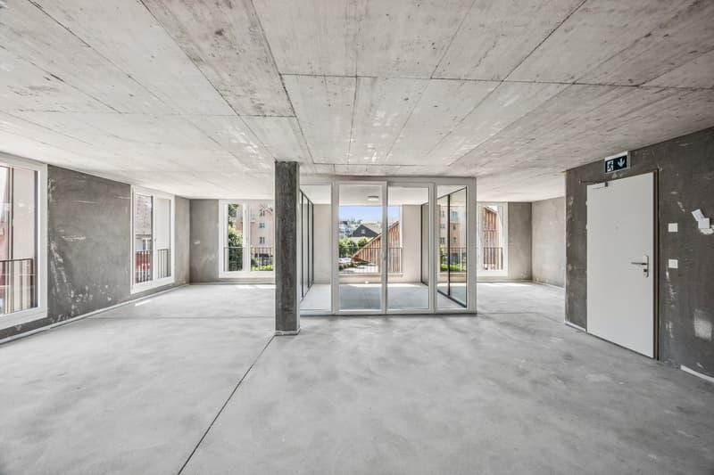 Large open space with concrete floors and ceiling, glass windows and doors, and minimal furnishings. The space appears to be a commercial or office area, ready for customization and renovation.