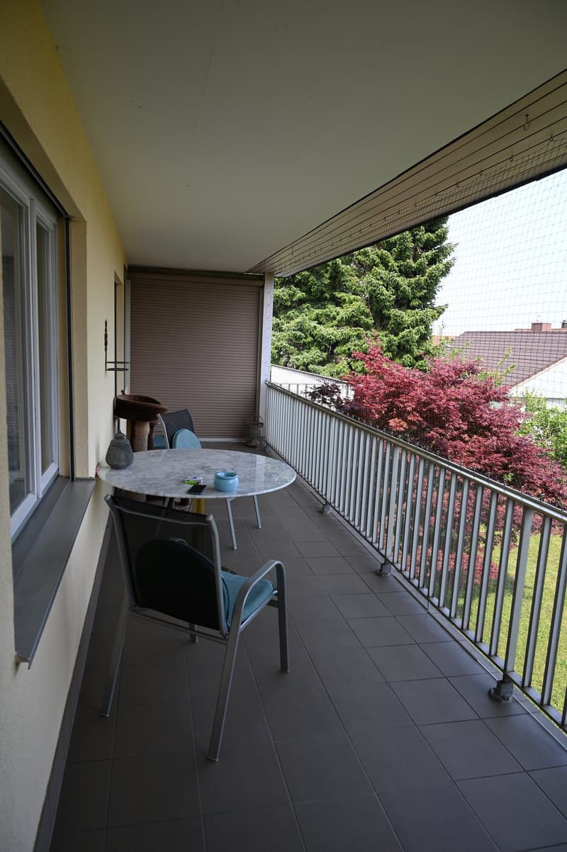 a covered balcony with tiled floor, metal railing, wooden chair, round table, and bird feeder; overlooking a garden with red leaves and a distant house