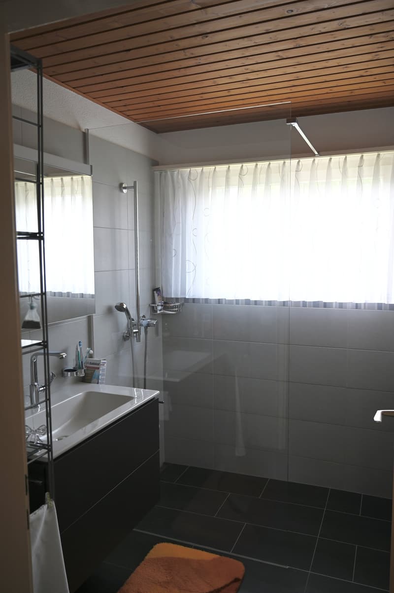 Modern bathroom, white tiled walls, white tiles on floor, white bathtub, sink with cabinet, mirror, shower with showerhead, window with curtains, wooden ceiling, toiletries
