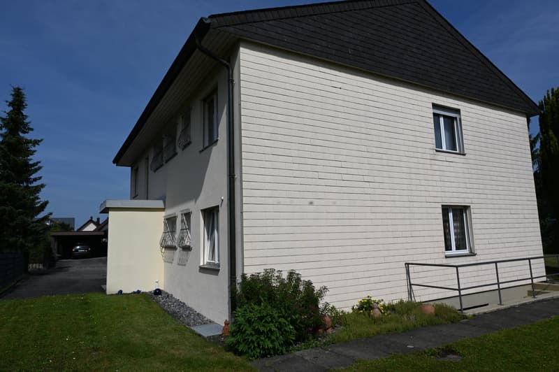 two story house, white brick wall, black roof, attached garage, metal railing, driveway, small plants in front