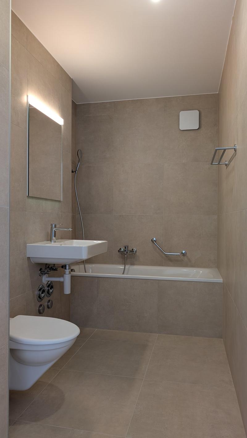 Bathroom with beige tiles, white sink, wall-mounted toilet, bathtub, mirror, showerhead, and wall-mounted towel rack. A power outlet is on the wall above the sink.