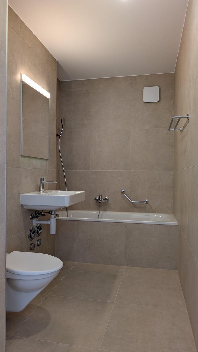 A bathroom with a white wall, white sink, bathtub, and toilet. A mirror, a faucet, and a handrail can be seen on the wall.