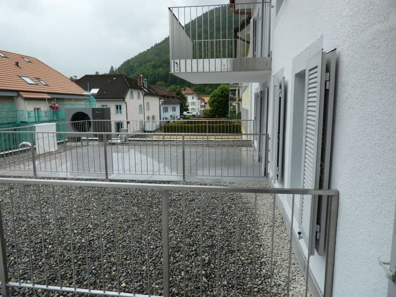 The image shows a multi-story apartment building with a balcony or terrace visible. The building has a white exterior and is surrounded by gravel and other residential buildings in a mountainous area.