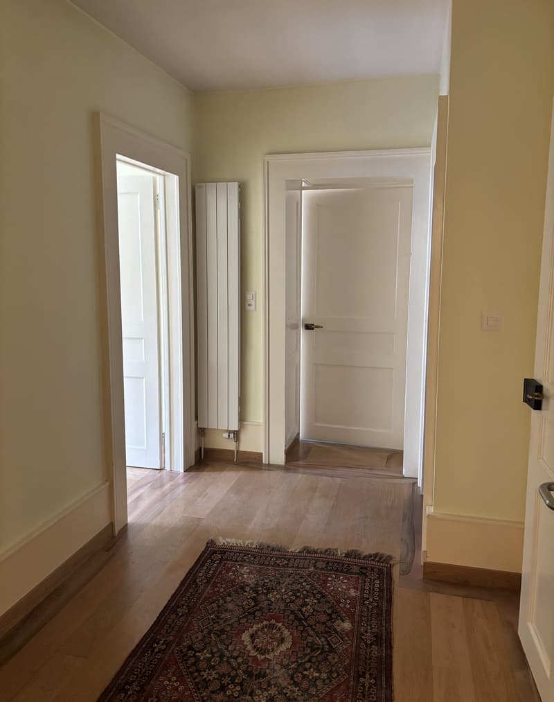 A long corridor with white walls, hardwood floors, two open doors leading to different rooms, a white radiator, and a rug in the middle.