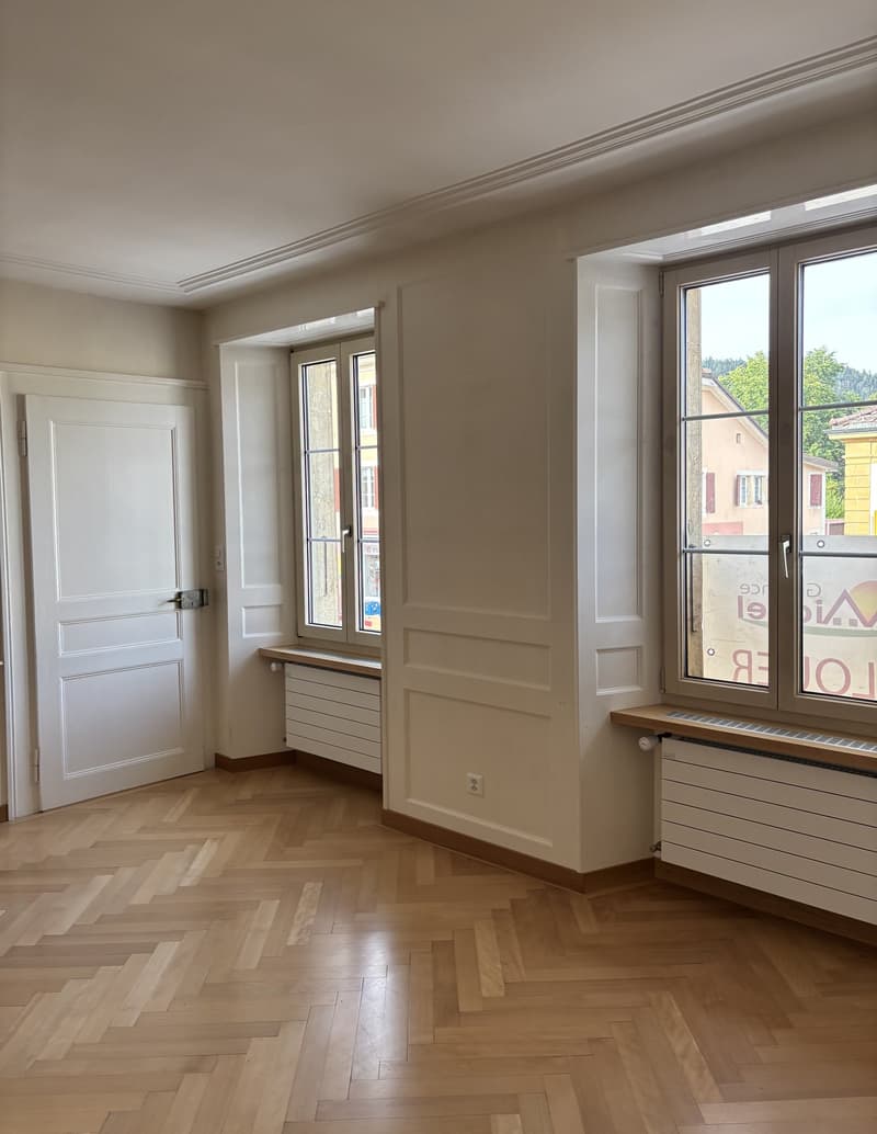 An empty room with white walls, wooden herringbone floor, a white door, double windows, and radiators under the windows. One of the windows provides a view of the buildings outside.
