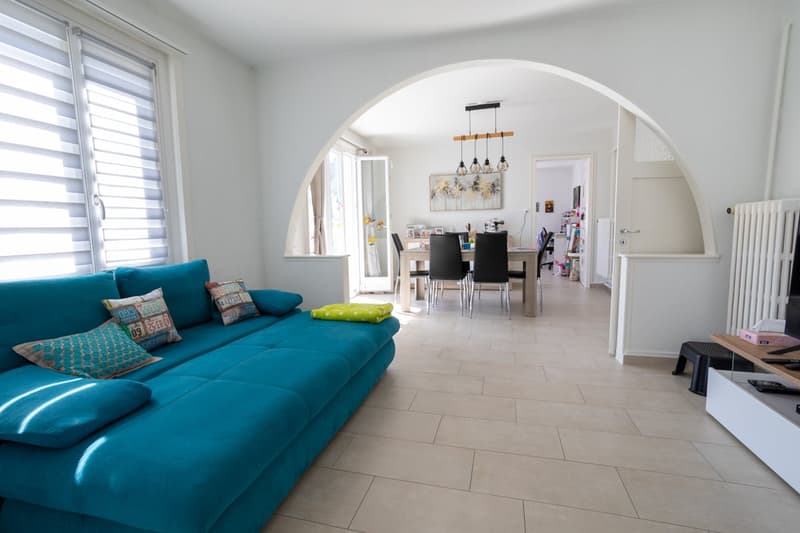 Living room with a blue sofa, decorative pillows, a dining table with chairs, and a chandelier