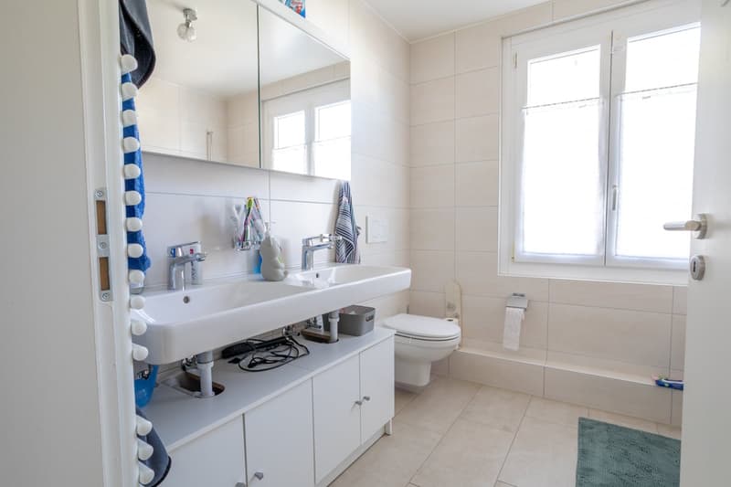 Bathroom with white tiles, double sink, toilet, mirror, and window