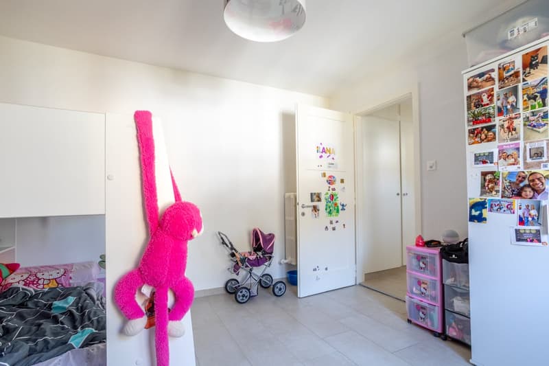 A room with a bed, a pink stuffed monkey, a stroller, a white door, a white refrigerator, and a white cabinet with pictures.