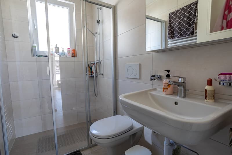 white tiles, glass shower, white sink, white toilet, white cabinet, mirror, toiletries