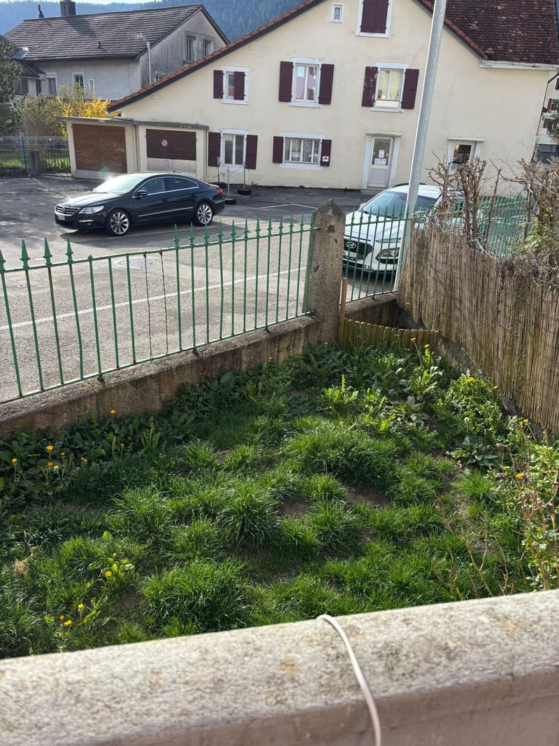 A small grassy area with a green metal fence and a wooden gate. There are two cars parked in front of a house with a brown roof and white walls. The house has several windows and a door. There are also some trees and plants in the area.