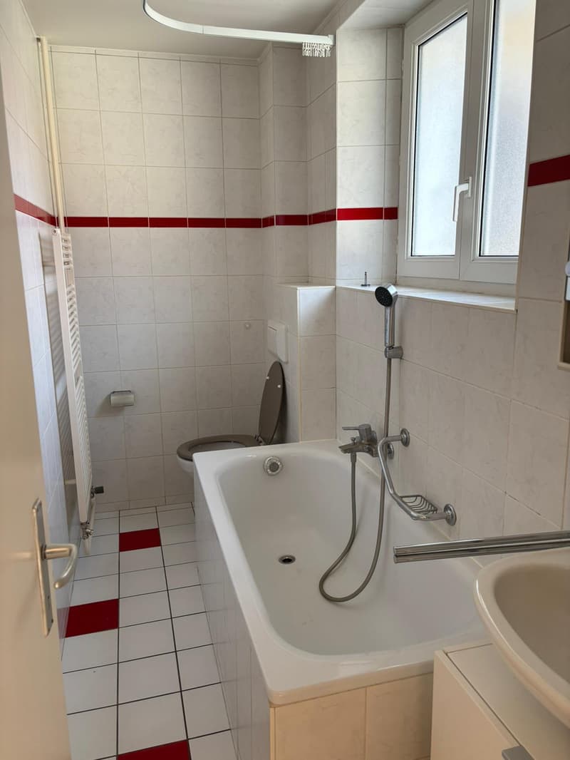 Bathroom with white tiles, red trim, bathtub, shower head, faucet, toilet, sink, and towel rack.