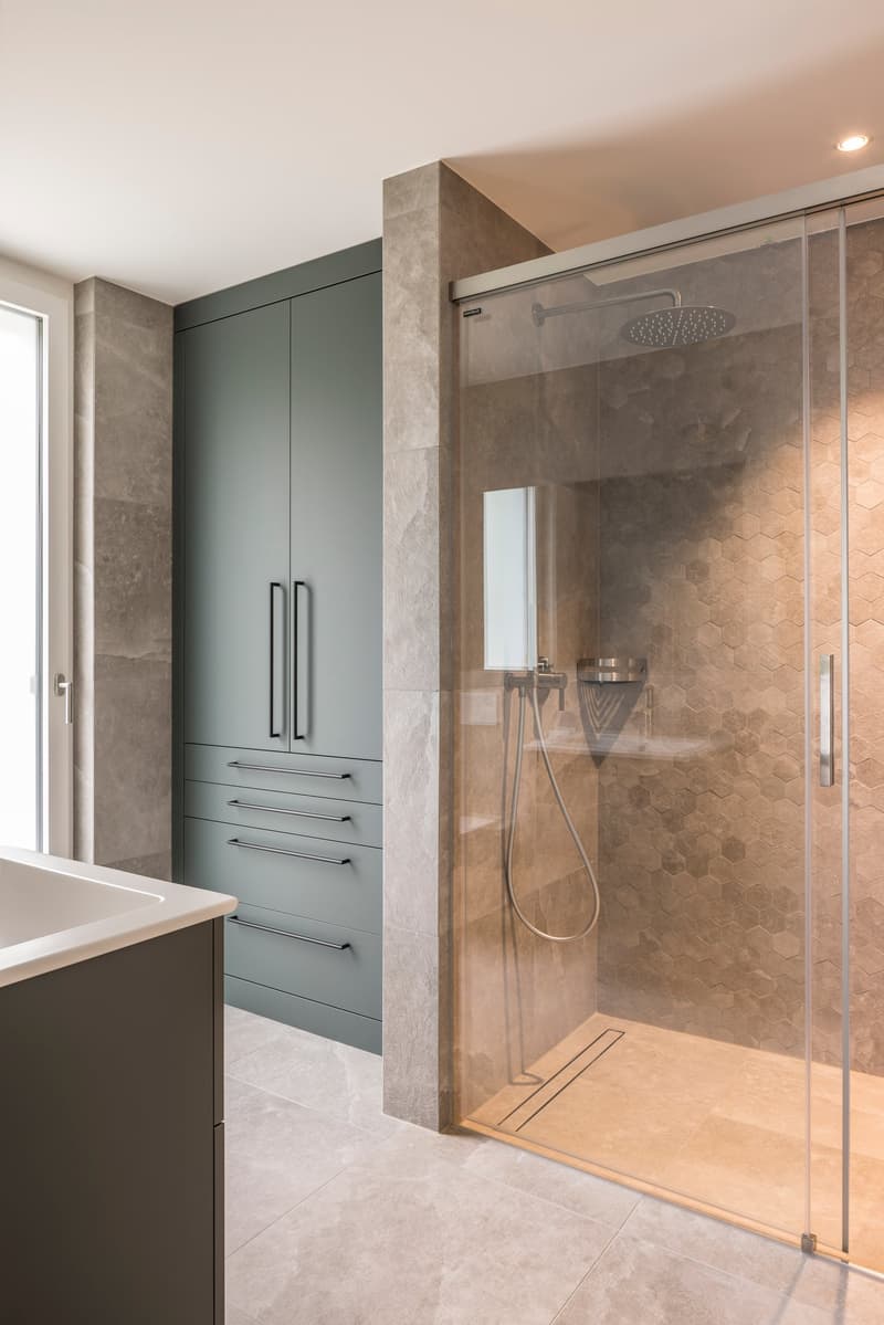 Bathroom with a glass shower enclosure, a sink, and a large mirror. The walls and floor are tiled with light-colored tiles. There is a large green cabinet with drawers and a door.