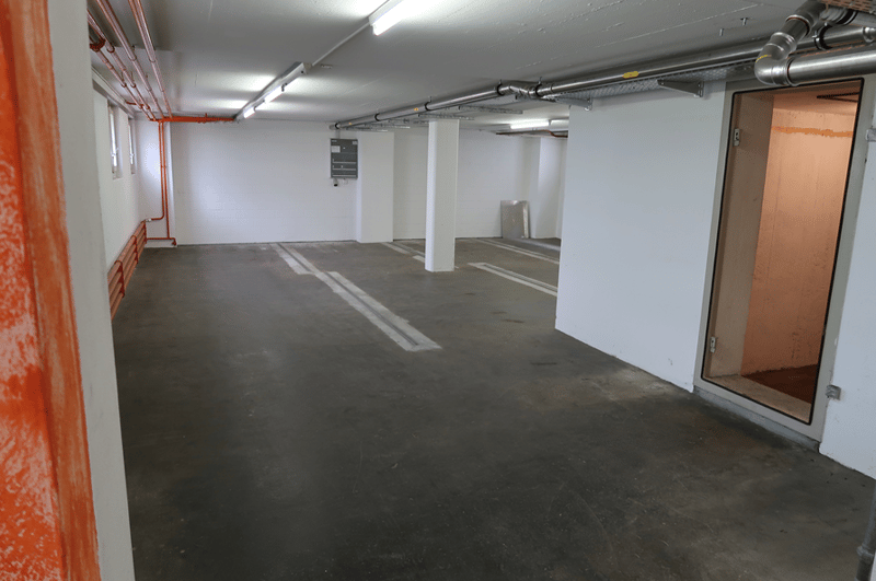 underground parking space, empty, white walls, ceiling with metal pipes, several parking spots with white markings, open doorway leading to a staircase