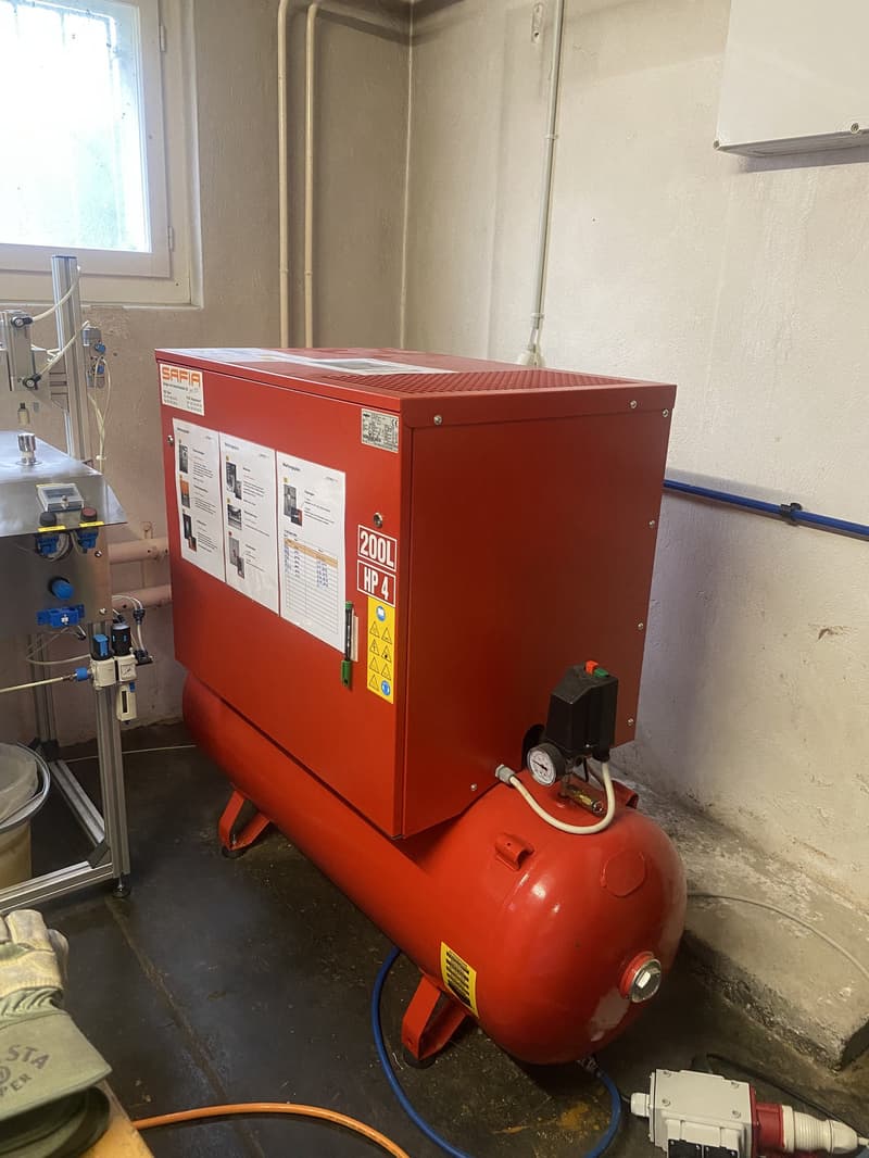 Large red compressor unit with model number 200L HP 4. Connected to pipes and electrical outlets, equipped with gauges and valves. Located in a utility room with white walls, window, and metallic structure.