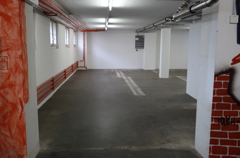 An empty basement parking area with concrete flooring, metal pillars, white walls, and red pipes.