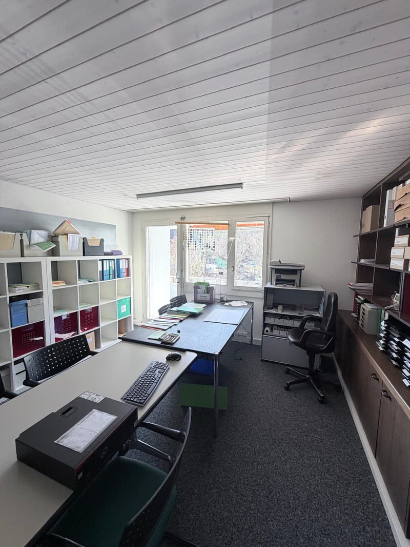 Long desk with computer, 3 black chairs, white shelves, grey carpet