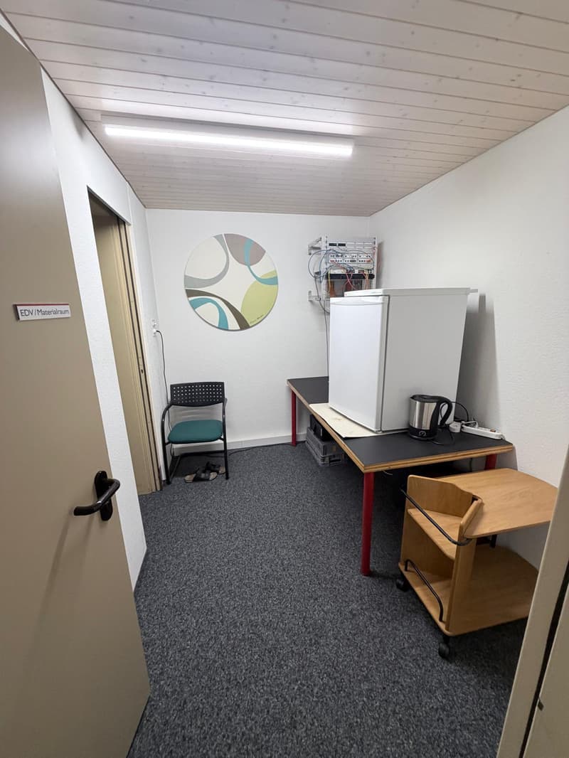 An empty room with a carpeted floor, white walls, ceiling with recessed lighting, a chair, a desk with a refrigerator, a kettle, and a shelf on wheels. There is a round artwork on the wall and a door on the left side.