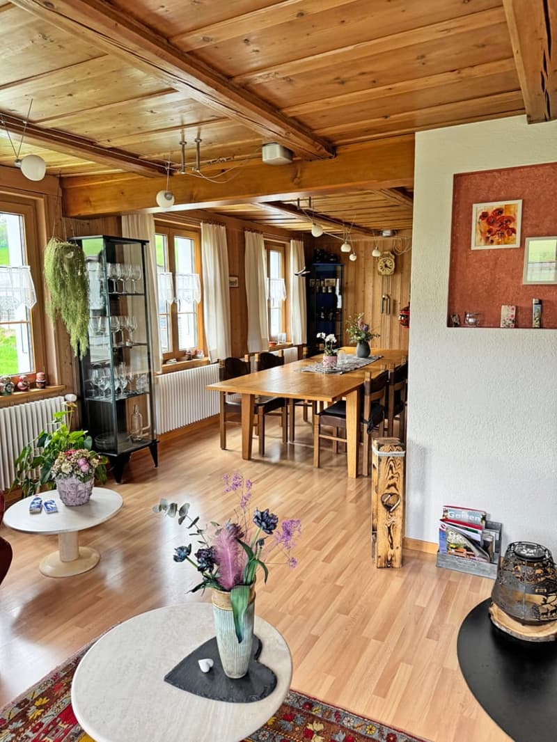 Dining room with wooden floor, wooden dining table, chairs, wooden ceiling, wooden shelves, radiator, windows, picture frame, flowers, plants, magazines, books